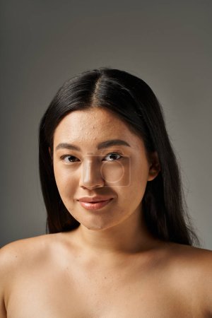happy young asian woman with skin issues and bare shoulders looking at camera on grey background