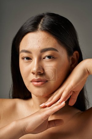 brünette junge asiatische Frau mit Hautproblemen und nackten Schultern schaut in die Kamera auf grauem Hintergrund