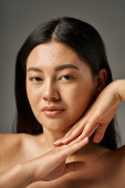 brunette young asian woman with skin issues and bare shoulders looking at camera on grey background Poster #692760466