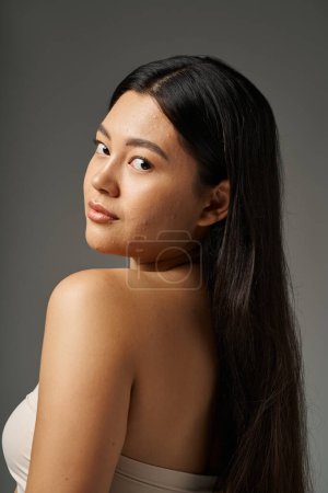 junge asiatische Frau mit brünetten Haaren und zu Akne neigender Haut beim Anblick des Spiegels im Badezimmer, Banner