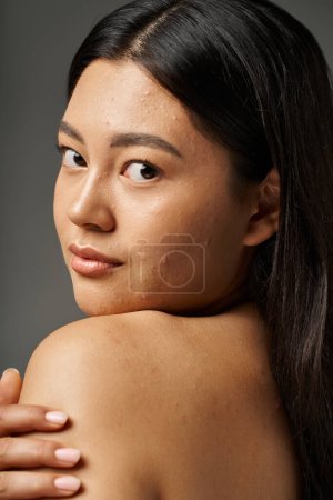 mujer asiática joven con pelo morena y piel propensa al acné mirando el espejo en el baño, pancarta