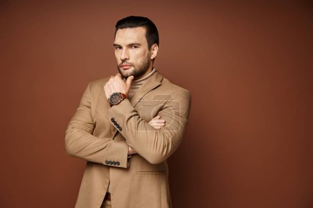 hombre guapo en traje elegante dando mirada confiada mientras posando con la mano cerca de la barbilla en beige