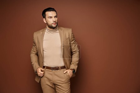 hombre guapo en traje elegante mirando hacia otro lado mientras posando con la mano en el bolsillo sobre fondo beige