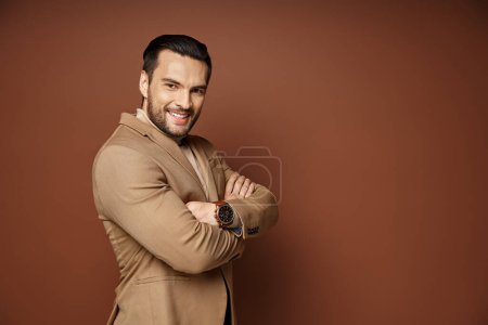 cheerful man with bristle posing in stylish beige blazer with folded arms on beige backdrop