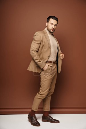 homme suave en tenue élégante regardant loin tout en posant avec la main dans la poche sur fond beige