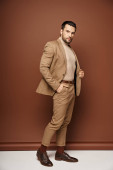suave man in elegant attire looking away while posing with hand in pocket on beige backdrop Poster #692773406