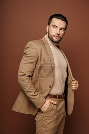 handsome man in elegant attire looking away while posing with hand in pocket on beige background