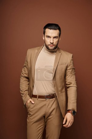 hombre guapo en traje elegante mirando a la cámara mientras posando con la mano en el bolsillo sobre fondo beige