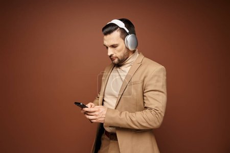 Foto de Hombre guapo en traje elegante usando su teléfono inteligente con auriculares, fondo beige - Imagen libre de derechos