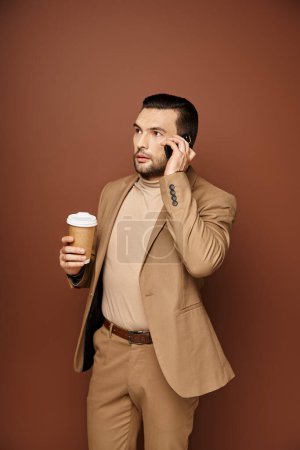 handsome man in stylish attire holding coffee to go and having call on smartphone on beige backdrop