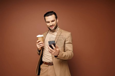 Téléchargez les photos : Homme heureux en tenue élégante tenant café pour aller et en utilisant son smartphone sur fond beige - en image libre de droit