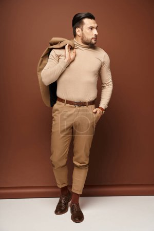 handsome man in turtleneck holding his jacket over shoulder and posing with hand in pocket on beige
