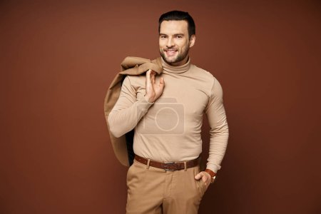 happy man in turtleneck holding his jacket over shoulder and posing with hand in pocket on beige