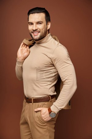 hombre feliz con cuello alto sosteniendo chaqueta de moda sobre el hombro y posando con la mano en el bolsillo en beige