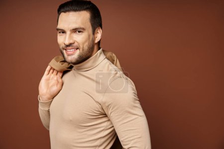 happy man in turtleneck holding trendy jacket over shoulder and looking at camera on beige