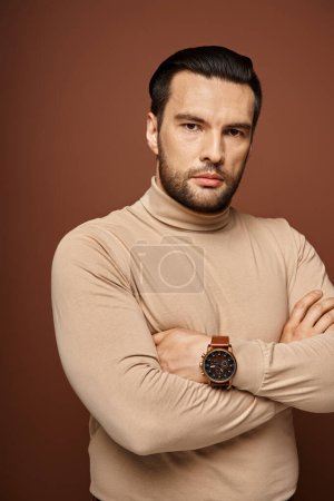 Hombre decidido y guapo con cuello alto posando con los brazos cruzados y la mirada aguda sobre fondo beige