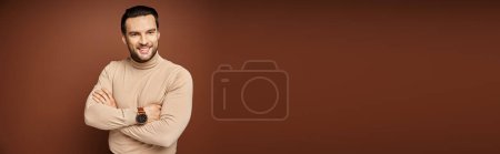 happy and handsome man in turtleneck posing with crossed arms on beige backdrop, banner