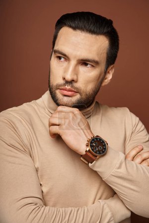 handsome man with bristle posing in turtleneck and touching his chin on beige background, male model