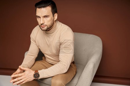 handsome and serious man in turtleneck sitting on comfortable armchair on beige background