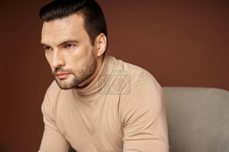 portrait of handsome and serious man in turtleneck sitting on armchair on beige background