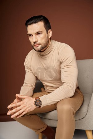 concentrated and serious man in turtleneck sitting on comfortable armchair on beige background
