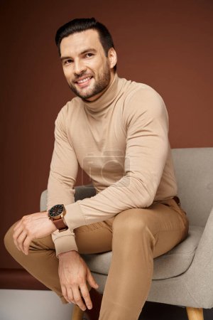 Photo for Positive and handsome man in turtleneck sitting on comfortable armchair on beige background - Royalty Free Image