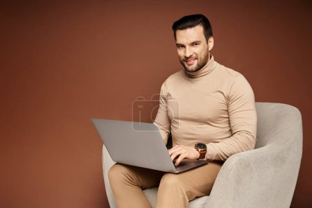Téléchargez les photos : Homme heureux à col roulé assis sur un fauteuil confortable et en utilisant un ordinateur portable sur fond beige - en image libre de droit