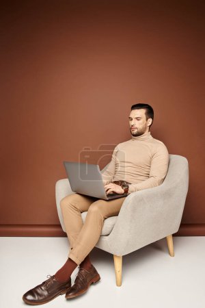 gut aussehender Mann im Rollkragen sitzt auf bequemen Sesseln und benutzt Laptop auf beigem Hintergrund