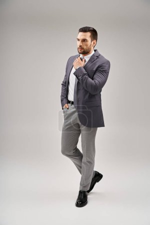 handsome man in sharp and stylish suit adjusting his tie and posing with hand in pocket on grey