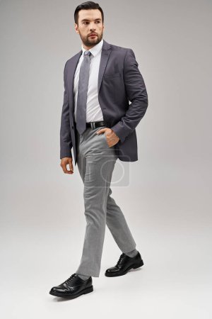 schöner Mann in scharfem und stilvollem Anzug, der mit der Hand in der Tasche auf grauem Hintergrund posiert, elegant