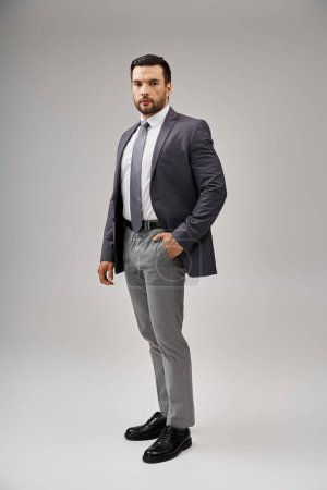 hombre guapo en traje afilado y elegante posando con la mano en el bolsillo sobre fondo gris, elegancia