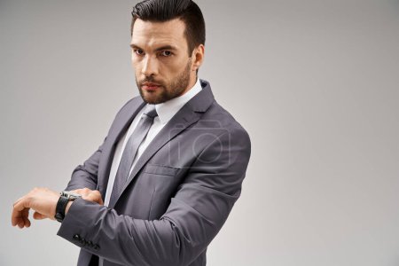 schöner Geschäftsmann posiert im eleganten Anzug und überprüft seine Armbanduhr auf grauem Hintergrund, Karriere