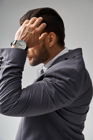 hombre guapo en ropa formal con reloj de pulsera ajustando su cabello sobre fondo gris, moda corporativa
