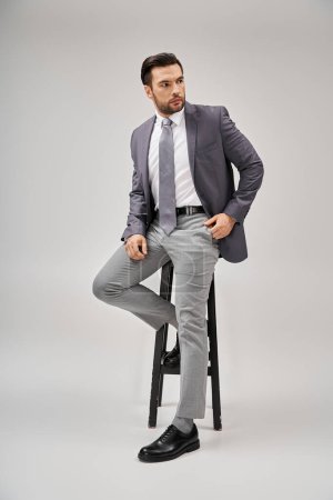 gutaussehender und selbstbewusster Geschäftsmann in formeller Kleidung, der auf einem hohen Hocker auf grauem Hintergrund lehnt