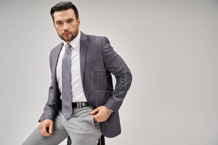 Téléchargez les photos : Homme d'affaires beau et confiant en tenue élégante assis sur un tabouret haut sur fond gris - en image libre de droit