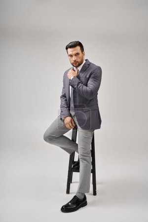 handsome and confident businessman in formal wear leaning on high stool on grey background, elegance