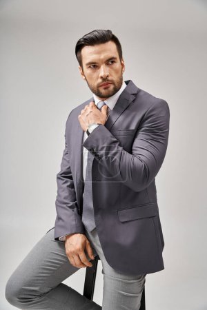 Téléchargez les photos : Homme d'affaires beau et confiant en tenue formelle appuyé sur un tabouret haut sur fond gris - en image libre de droit