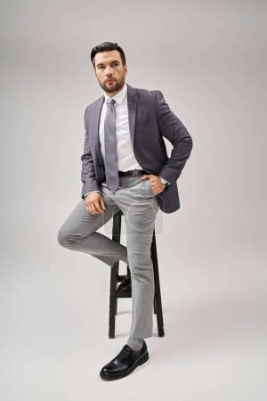 Photo for Handsome businessman with bristle posing with hand in pocket in suit and sitting on high stool - Royalty Free Image