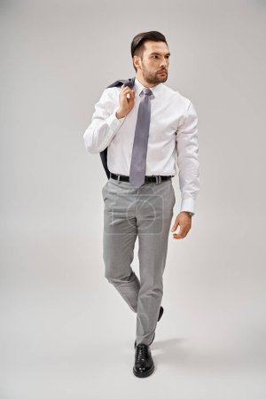 businessman in formal wear holding jacket over shoulder while walking on grey background