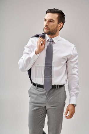 Geschäftsmann in seinen Dreißigern mit Borstenjacke über der Schulter, während er auf grauem Hintergrund steht