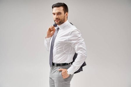 hombre de negocios con cerdas sosteniendo la chaqueta sobre el hombro mientras está de pie con la mano en el bolsillo en gris