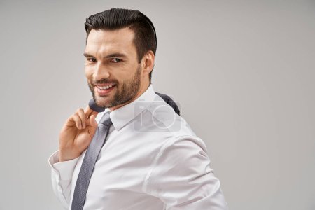 Porträt eines glücklichen Geschäftsmannes in seinen 30ern, der Jacke über der Schulter hält, während er auf grau steht