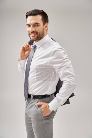 hombre de negocios sonriente con cerdas sosteniendo la chaqueta sobre el hombro mientras está de pie con la mano en el bolsillo