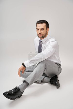 Foto de Hombre de negocios guapo en ropa formal elegante sentado en el suelo en el estudio con fondo gris - Imagen libre de derechos