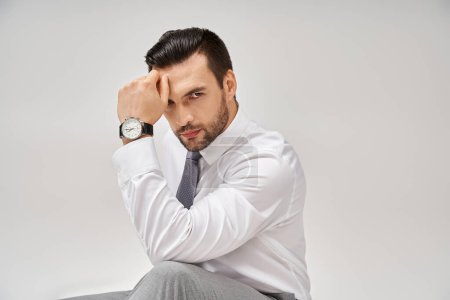 Téléchargez les photos : Homme d'affaires bien habillé et beau en tenue formelle assis sur le sol dans un studio avec fond gris - en image libre de droit
