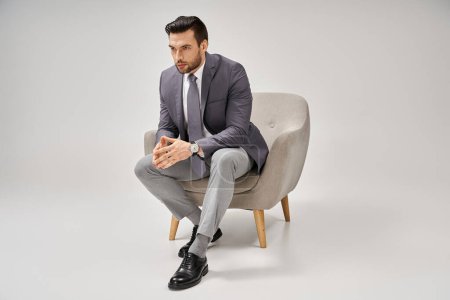 Foto de Hombre de negocios seguro y guapo en traje elegante sentado en sillón cómodo sobre fondo gris - Imagen libre de derechos