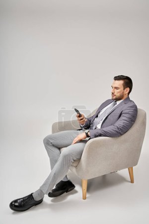 relajado hombre de negocios sentado en el sillón y el uso de teléfono inteligente sobre fondo gris, redes sociales