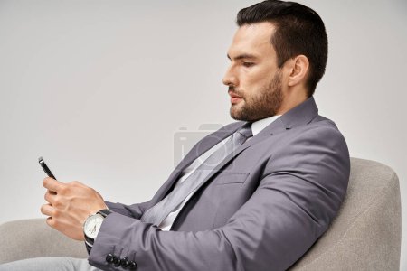 businessman with bristle sitting on armchair and using smartphone on grey background, social media