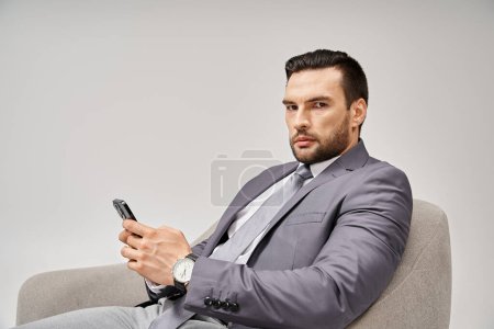 businessman with bristle sitting on armchair and using smartphone on grey background, connected