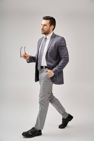 confident businessman in formal wear holding glasses and walking with hand in pocket on grey